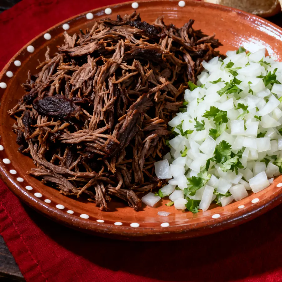 Shredded beef barbacoa next to chopped onion with cilantro on a clay plate