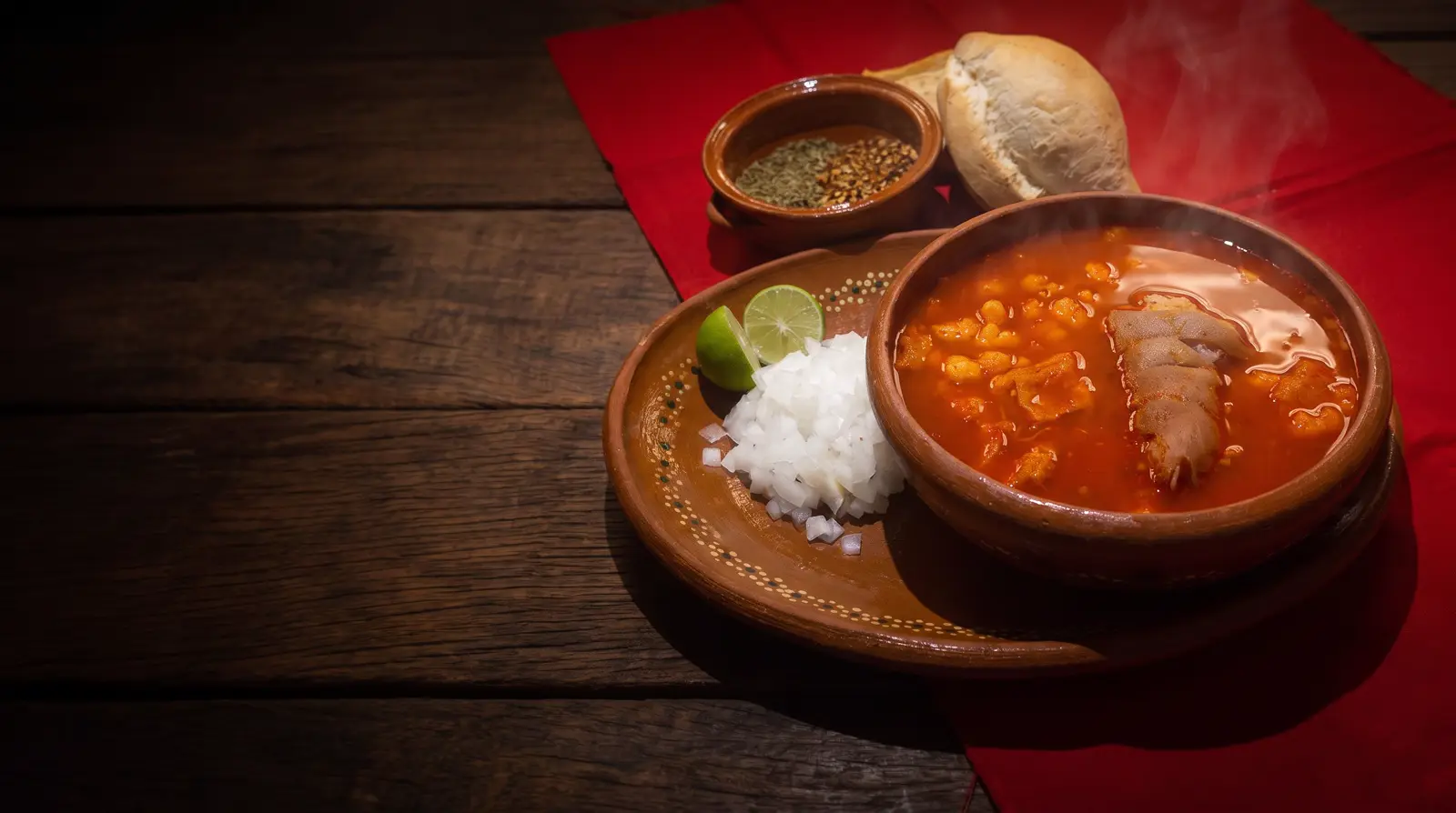 A clay bowl of red menudo with a plate of onion and lime, oregano, chile and a bolillo, on a red cloth
